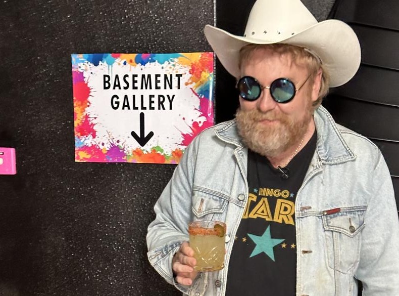 Luke Herbert in a cowboy hat beside the Basement Gallery sign